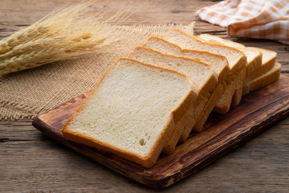 Four slices of white bread on a wooden chopping board, with strands of wheat in the background. loading=